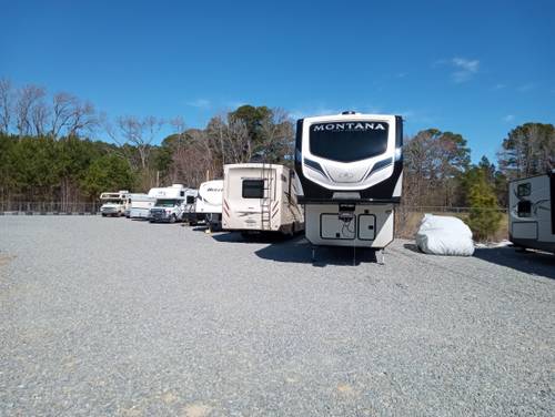 Large outdoor parking lot with various recreational vehicles, including fifth wheels and motorhomes, on a gravel surface under a clear blue sky.