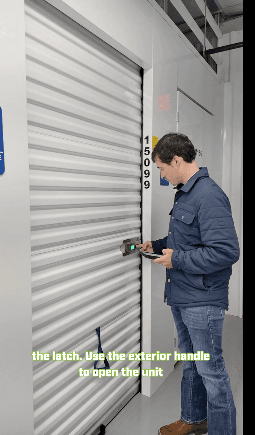 A man in a blue jacket and jeans using a keypad on a large, gray storage unit with a white roll-up door, next to a door with a yellow label.