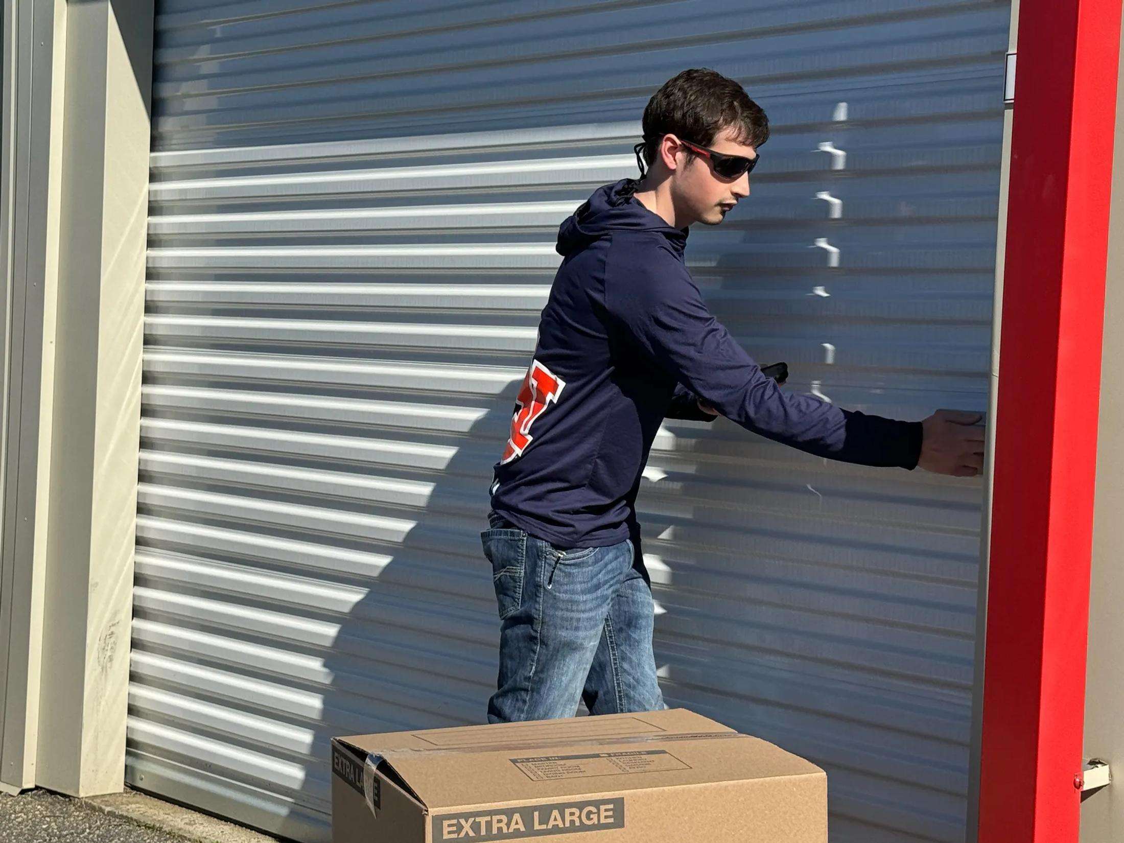 A young man wearing sunglasses and a navy hoodie is closing a metal storage unit door, with a cardboard box labeled "Extra Large" on the ground nearby.