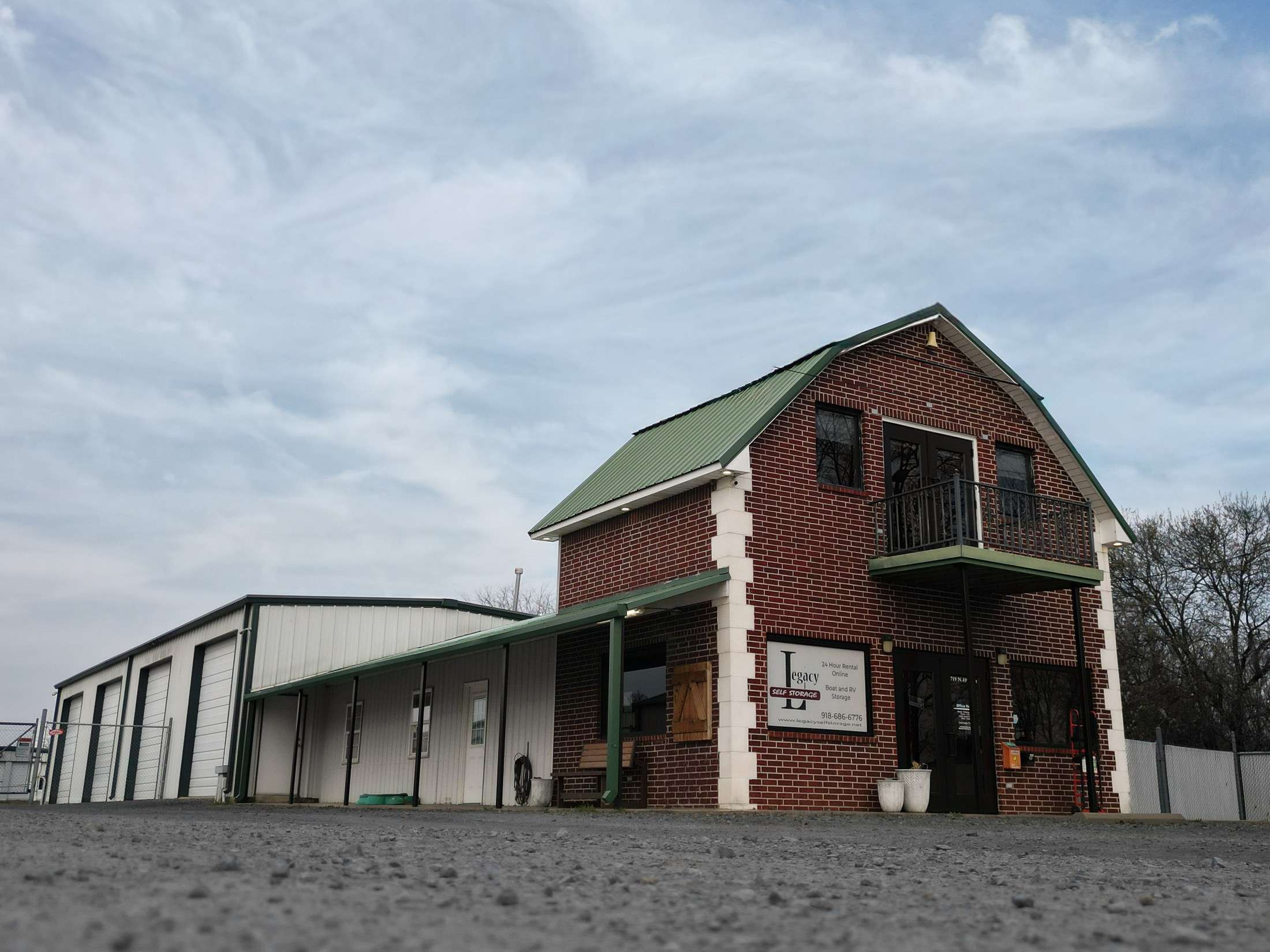 A two-story brick building with a small balcony and a green metal roof, serving as a storage facility with signs indicating "Legacy" storage services.