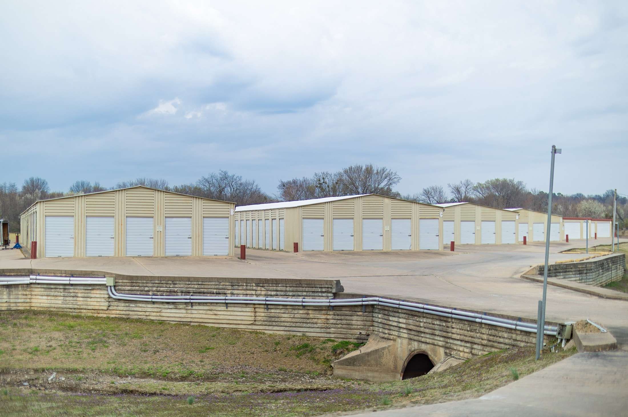 Storage units with metal roll-up doors on a concrete lot, surrounded by a retaining wall and utility pipes, under a cloudy sky.