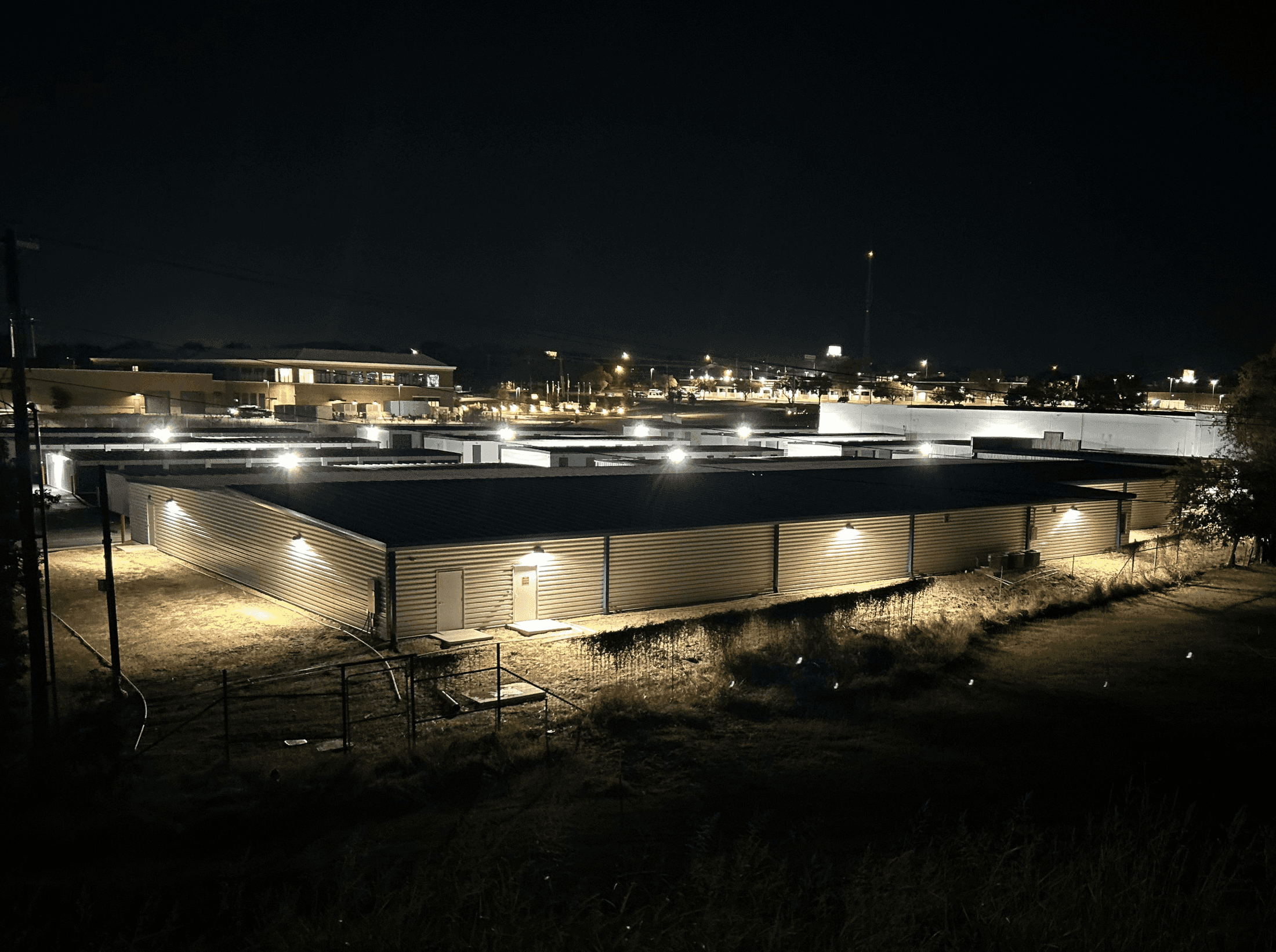 Steel storage units with exterior lighting at night, surrounded by a chain-link fence, in a commercial storage facility.