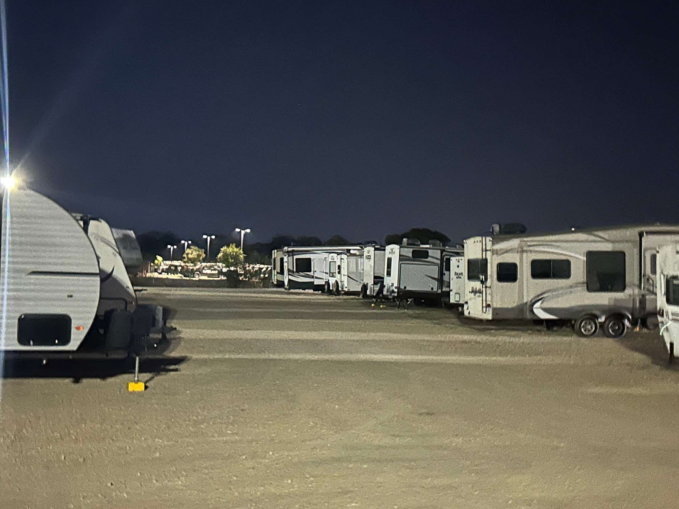 Line of travel trailers and RVs parked in a storage lot at night, illuminated by streetlights against a dark sky.