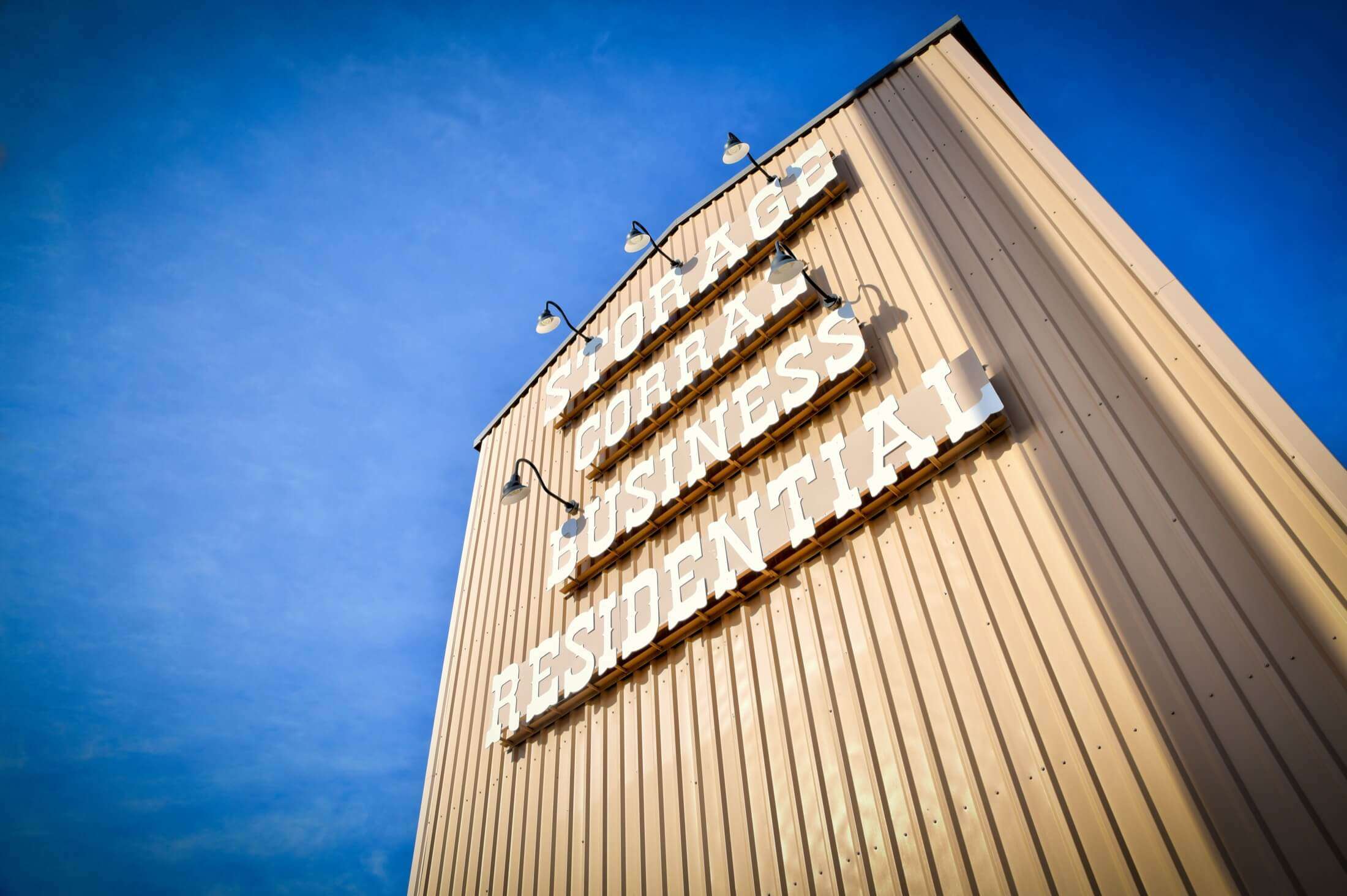 Storage business with beige metal exterior, large white signage reading "STORAGE," "CORRAL," "BUSINESS," and "RESIDENTIAL," and outdoor lighting fixtures against a blue sky.