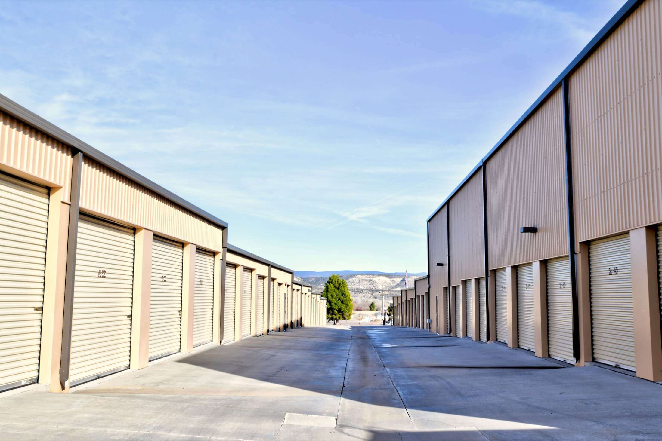 A spacious outdoor storage facility features multiple rows of storage units, each with beige roll-up doors. The asphalt driveway runs between the units, providing access to each door. The facility is well-maintained, with a clear blue sky overhead and distant mountains visible in the background.